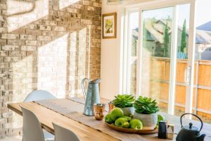 Sunlit dining room with contemporary decor and a rustic brick wall, featuring stylish lighting and a wooden table.